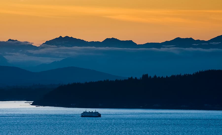 A Washington State Ferry sails past Bainbridge Island at sunset with the Olympic Mountains in the background.